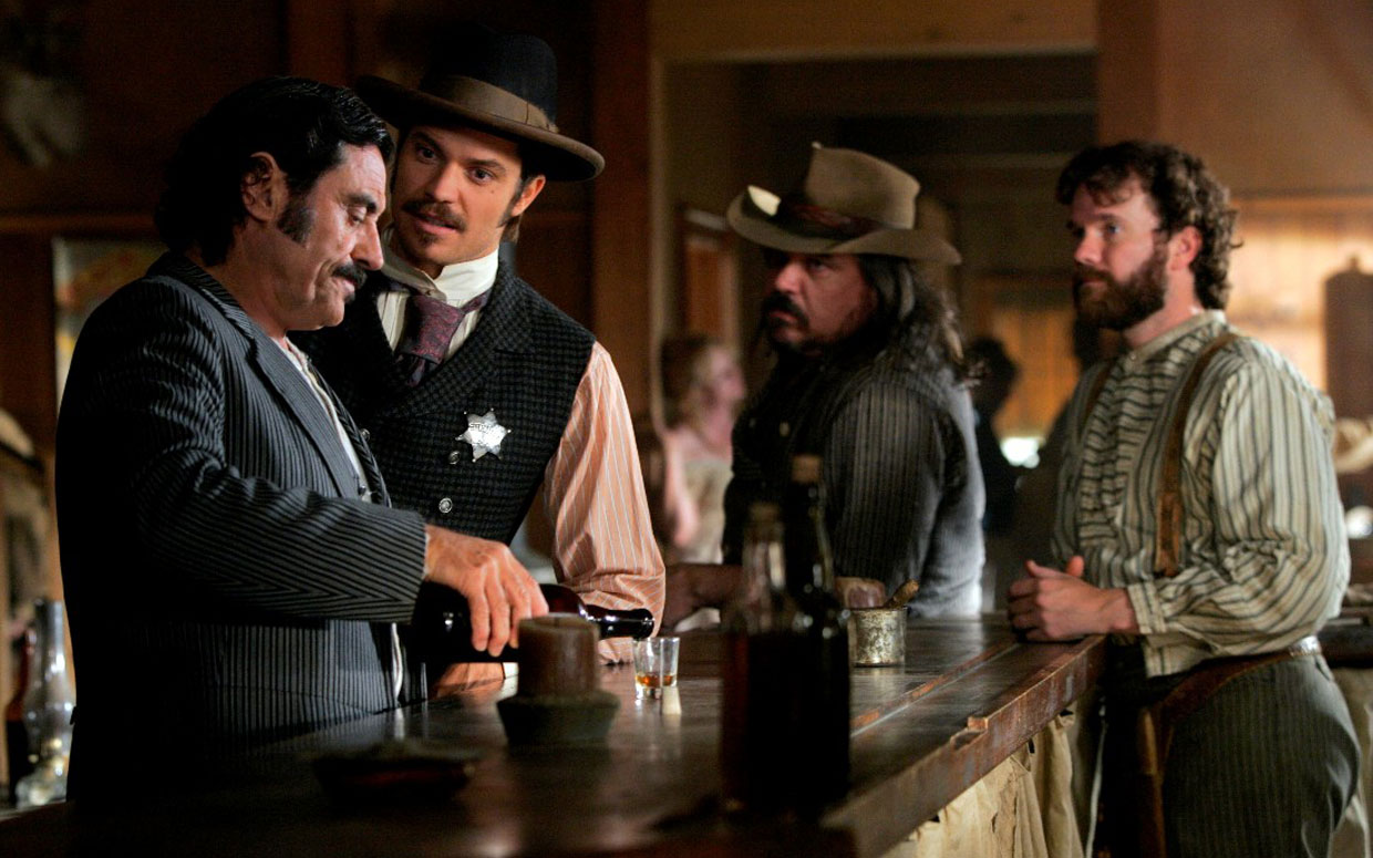 Vier MÃ¤nner in einem Saloon, ein Mann mit Sheriffstern redet ernst mit einem anderen, der gerade ein Glas Whisky eingieÃŸt.