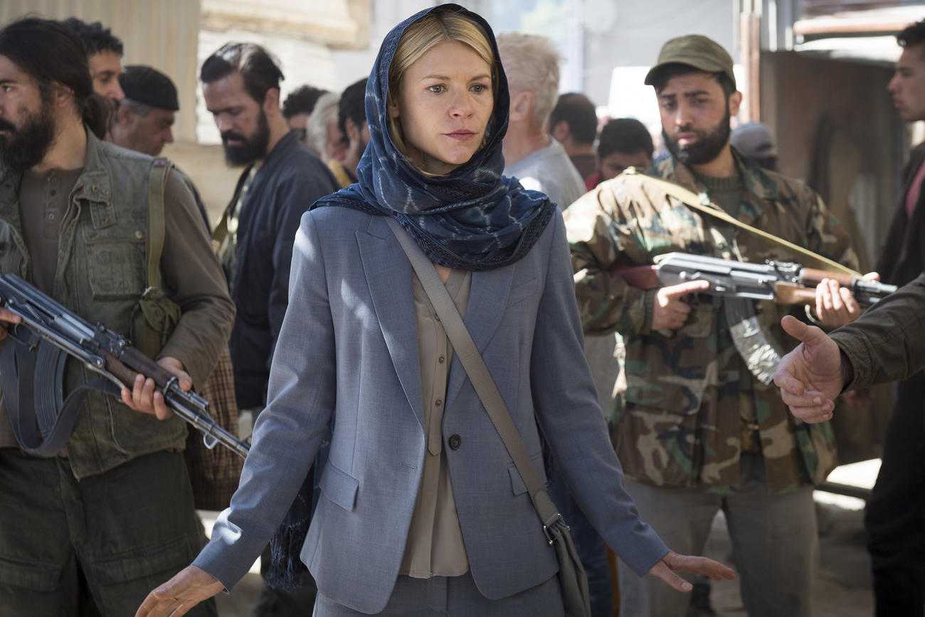 Eine blonde Frau mit Kopftuch und Hosenanzug, umgeben von Männern mit Waffen.